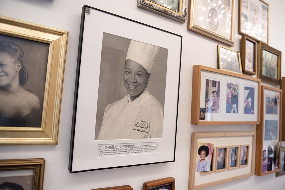 A wall of framed photographs in Chef Toni's home in Harlem, New York, centers a black and white image of her younger self in chef's hat and coat.