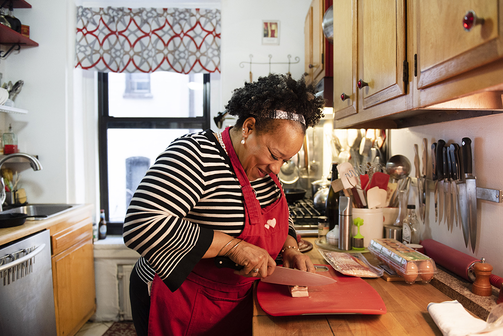 Chef Toni smiles over a red cutting board at her kitchen counter where she slices bacon, her red chef's apron protecting her black and white striped blouse, maple cabinets and a window looking out to apartment buildings beyond are visible in the background.