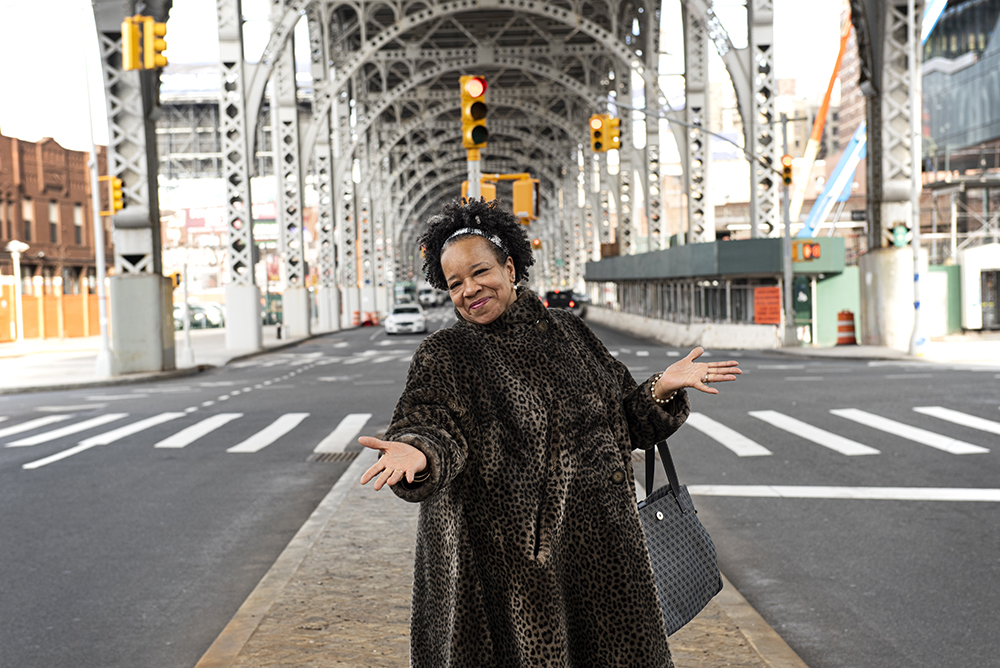 Standing in the median under the elevated highway, Chef Toni appears to be running the show that is New York City as she holds her hands wide to welcome you to her home. A black handbag rests near her elbow, and yellow traffic lights decorate the streets around her.