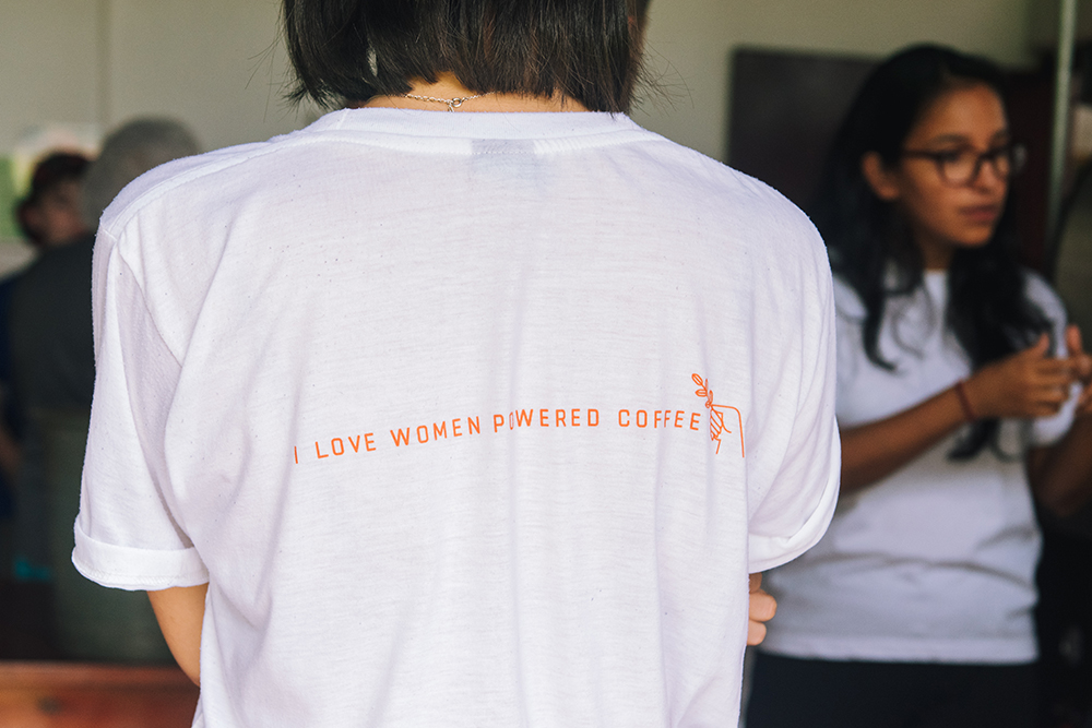A woman with short, straight dark hair is photographed from behind wearing a white t-shirt that reads "I love women powered coffee" in orange capital letters across her back. 