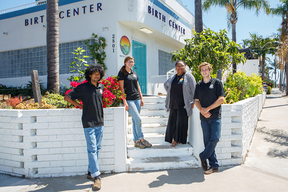 A bright white building with glass block panels and a turquoise door, large black letters reading 'birth center' on each side, and palm trees and tropical pink flowers lining the white brick beds in front of it. Employees of the San Diego Community Birth Center and San Diego Co Harvest, pose in front for a photo wearing black shirts and jeans.