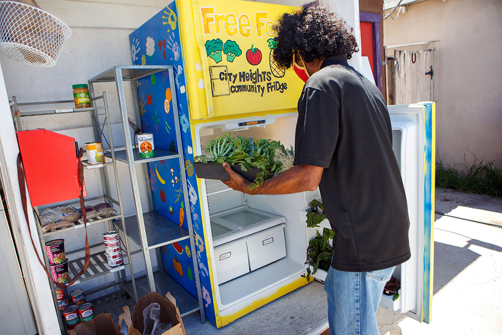 Marcus Slaughter loads a tray of green vegetables into a community refrigerator, painted by hand with images of fruits and vegetables on blue or yellow backgrounds. The words "city heights community fridge" can be seen on the freezer door.