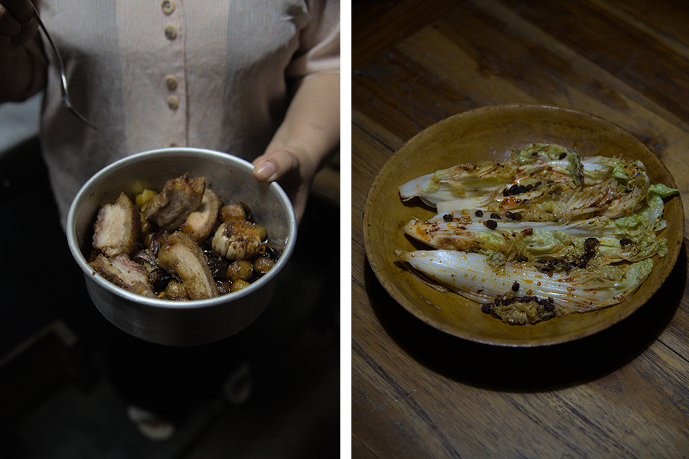 Zhimomi's mother holds a round stainless steel pan of roast pork belly up to the light for tasting, and next to that a wooden bowl of Chinese cabbage dressed with Axone and other spices sits atop a wooden dining table.