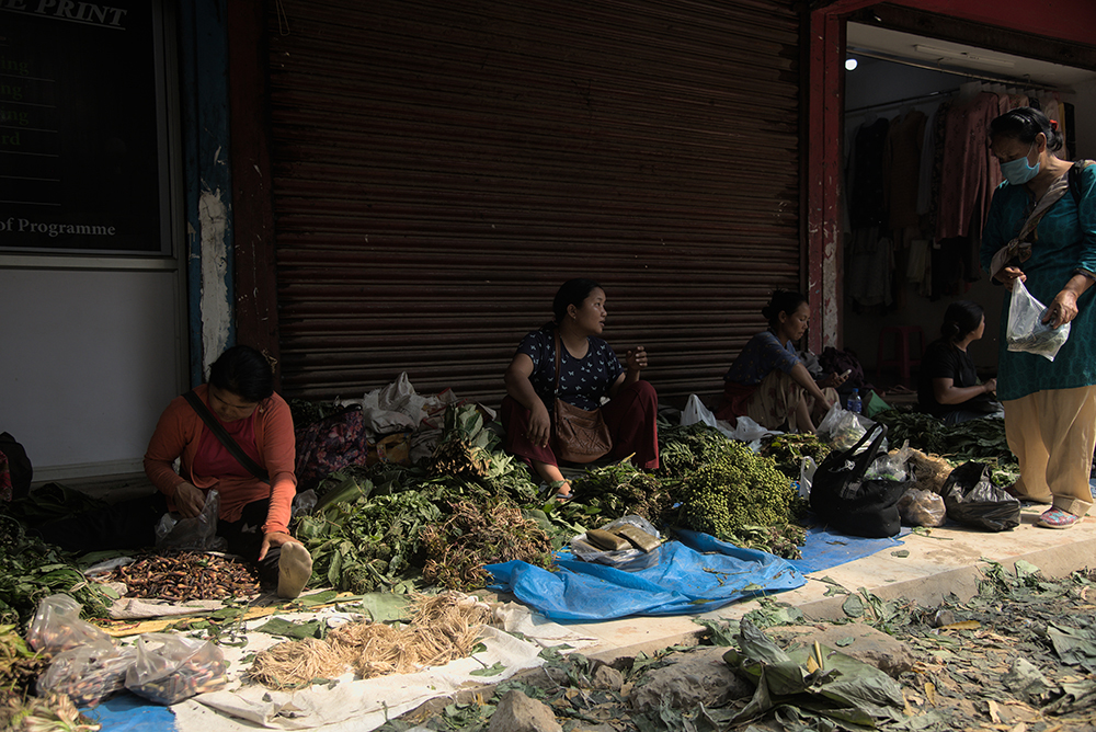 A line of three women sitting against the closed metal gate of a market stall with vegetables spread on tarps in front of them, speaking to a customer who stands to the right wearing a mask and holding a plastic bag of items purchased at the market.