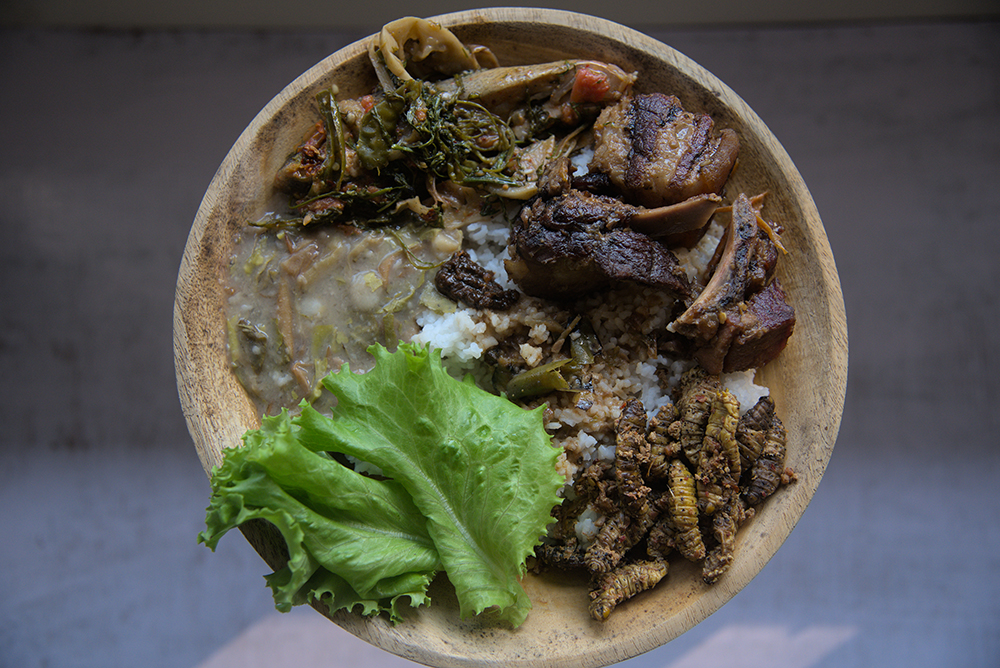 A wooden bowl holds smoked pork cooked with Axone, rice, cooked greens, and a bright green lettuce leaf for garnish.