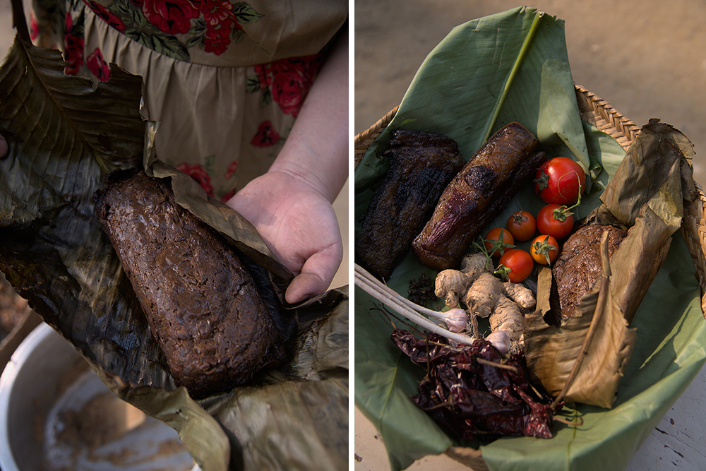 A woman unfolds the leaves encasing a completely fermented and smoked batch of dry, dark brown Axone. Next to her, a basket of ingredients used to make smoked pork with Axone, including tomatoes on the vine, garlic, ginger, and dried peppers.